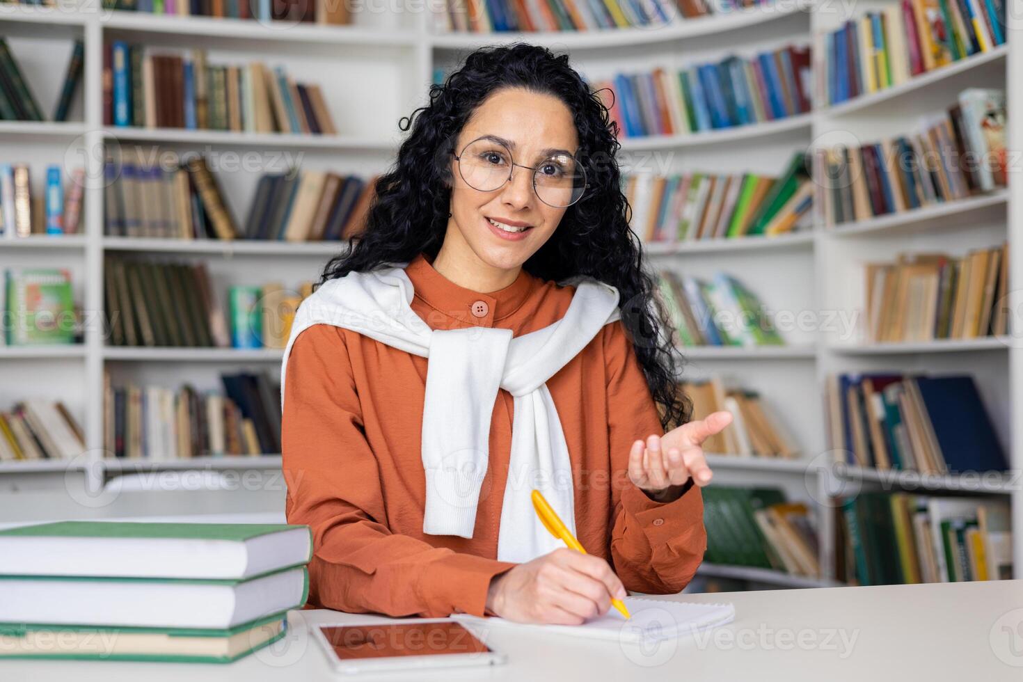 Young Hindu female teacher conducts online lesson in classroom sitting at the table, gesturing with hands and writing in notebook, camera view, office worker having work call looking at camera. photo