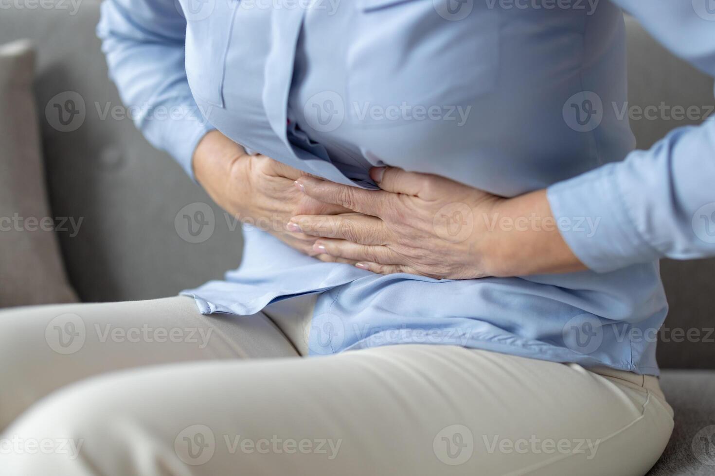 Close-up of a senior woman in pain, holding her abdomen while sitting on a sofa. The image depicts discomfort and the challenges of aging. photo