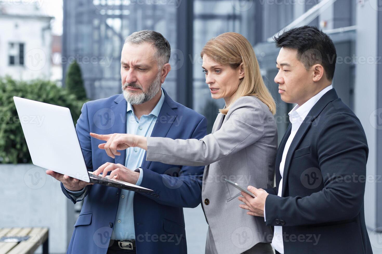 Successful business team, three colleagues businessman and businesswoman outside office building discussing current plans and management, looking at laptop screen, discussing and consulting photo