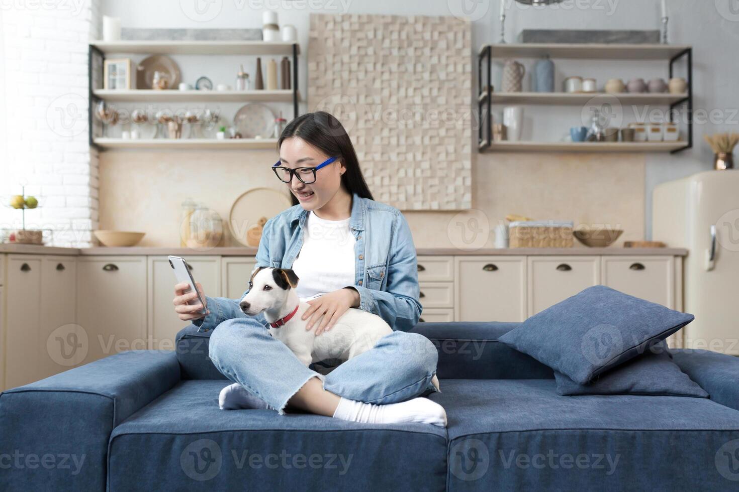 A happy young beautiful Asian woman in glasses is talking on a call, holding a phone and her dog in her hands, smiling at the camera. Sitting at home on the sofa in the lotus position. photo