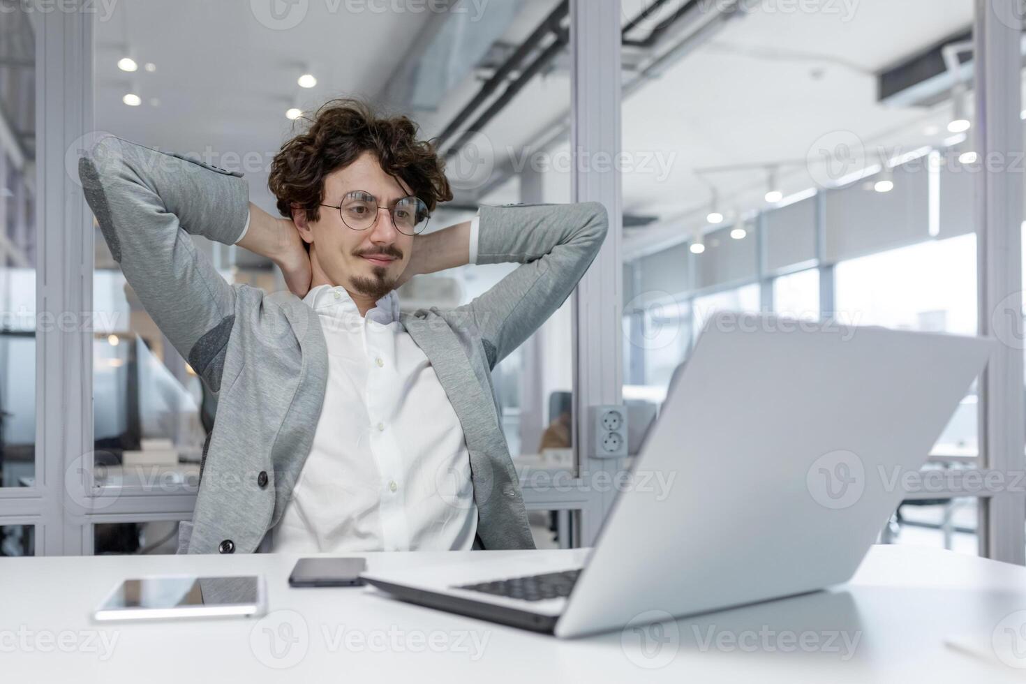 Young male entrepreneur with curly hair taking a break at his desk in a well-lit contemporary office, exuding confidence and professionalism. photo