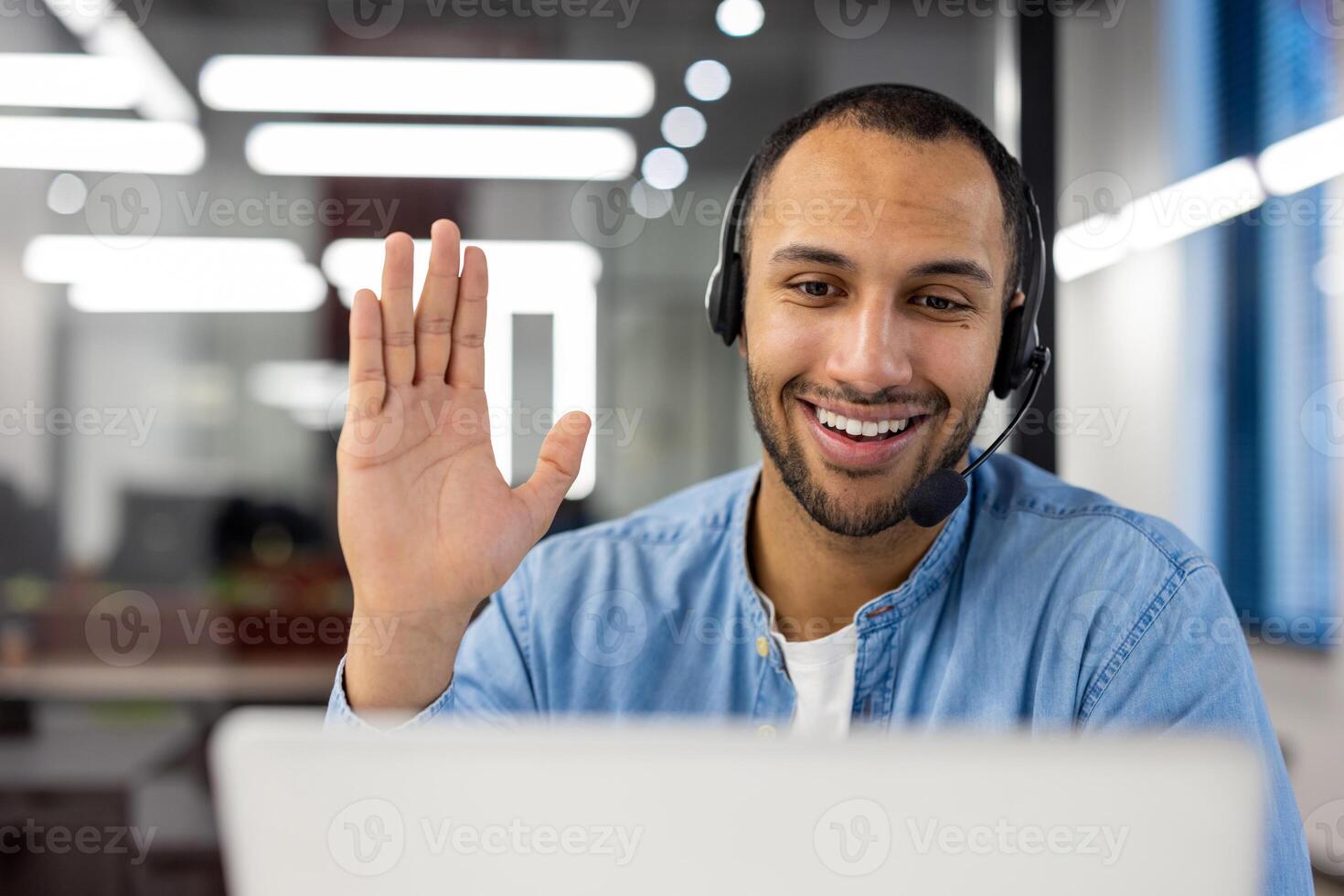 Close-up photo of a young smiling Arab man in a headset talking on a call on a laptop while waving his, hand at the camera.