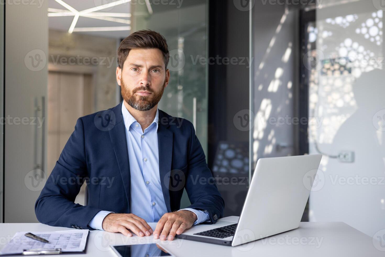 retrato de un exitoso y confidente joven hombre abogado en un negocio traje sentado a un oficina escritorio, trabajando con un ordenador portátil y documentos, mirando seriamente a el cámara. foto