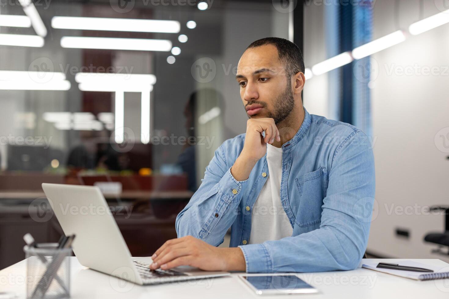 Pensive young hispanic man working and studying in office, sitting at desk holding head in hand and looking at laptop, analyzing data, looking for solution, thinking about project. photo