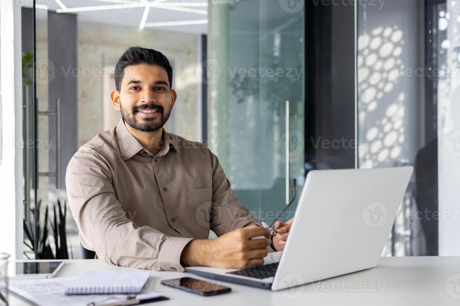 enfocado indio empresario utilizando un ordenador portátil a su escritorio en un brillante contemporáneo oficina configuración, con papeleo y digital tableta. foto