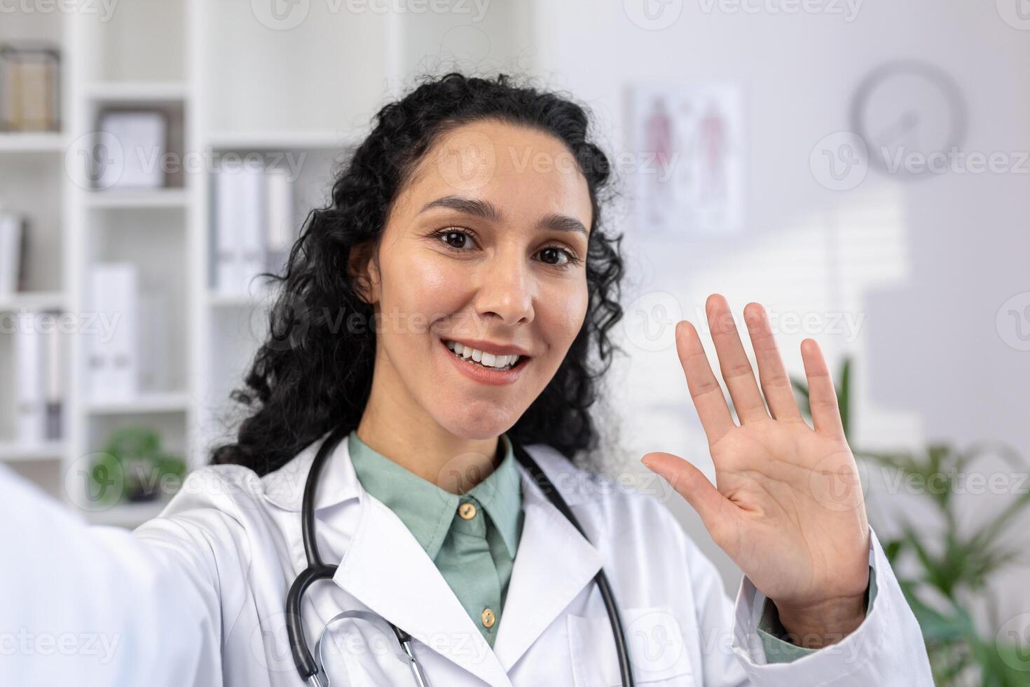 Female doctor smiling and looking at smartphone camera waving hand joyfully greeting, Hispanic woman in white medical coat talking using app on phone for call, standing inside clinic. photo