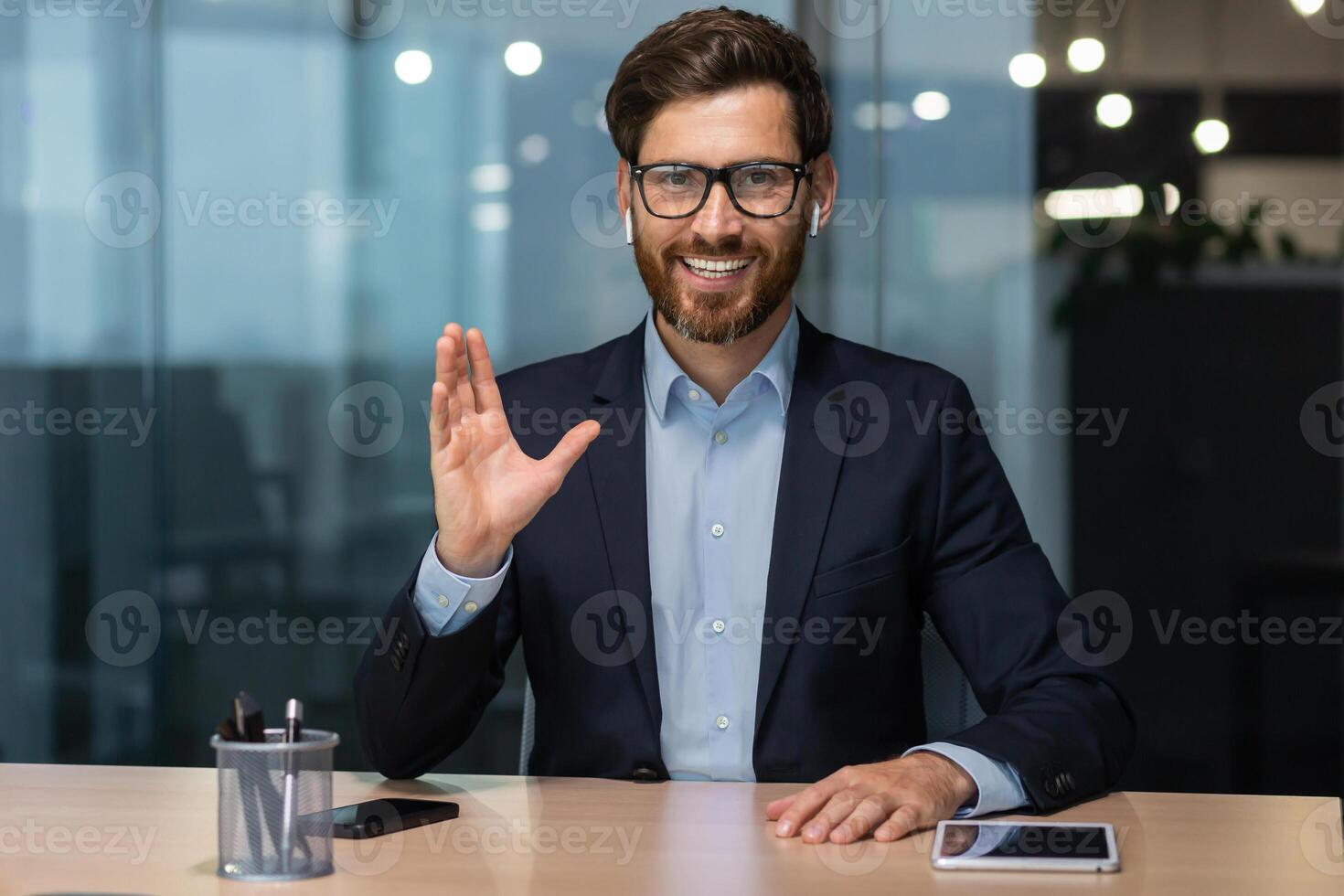 call on online meeting with colleagues, mature boss in business suit looking at web camera and smiling waving to employees, businessman in glasses working inside modern office building. photo