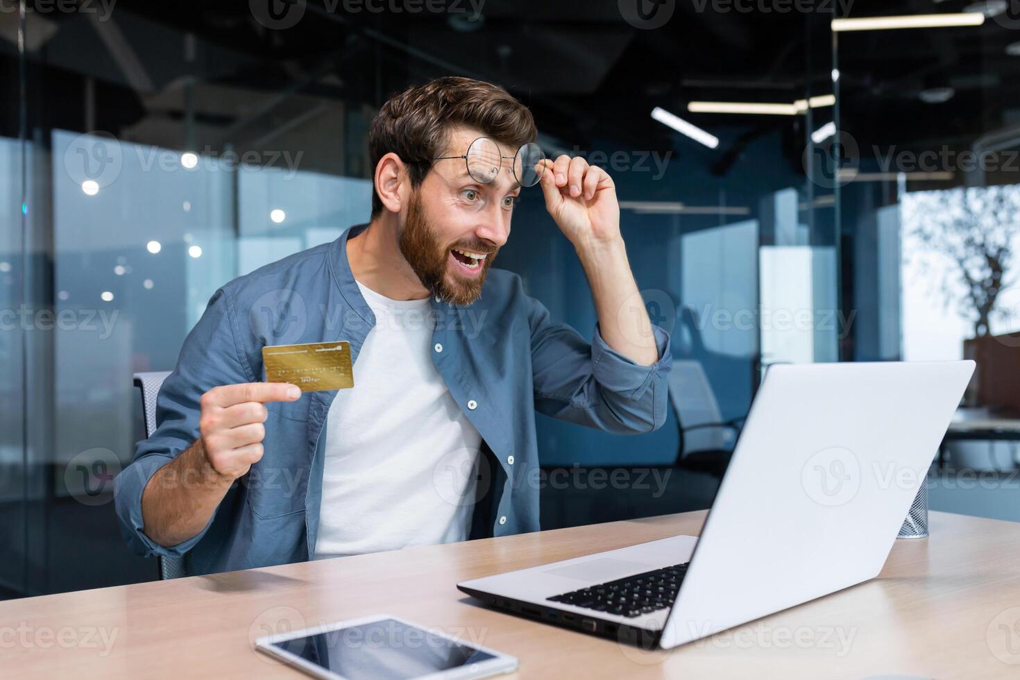 Cheated and disappointed businessman working inside office, man holding bank credit card and trying to make a purchase in online store, online fraud and deception. photo