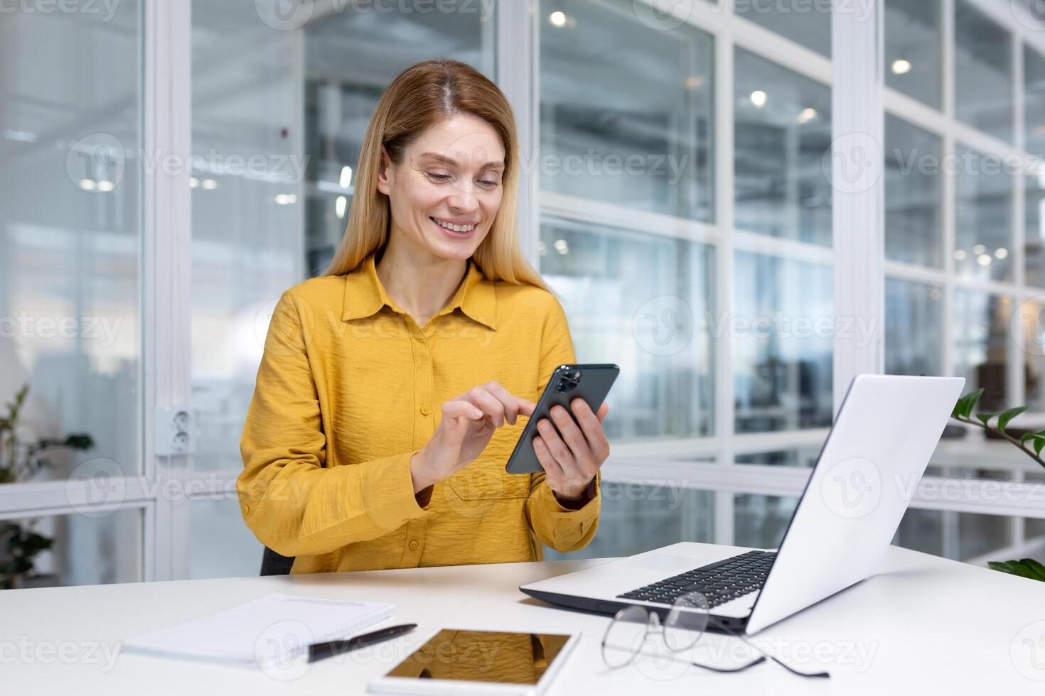 Mature experienced business woman inside office at workplace, employee sitting at desk smiling holding phone in hand, boss using application on smartphone, browsing internet pages. photo