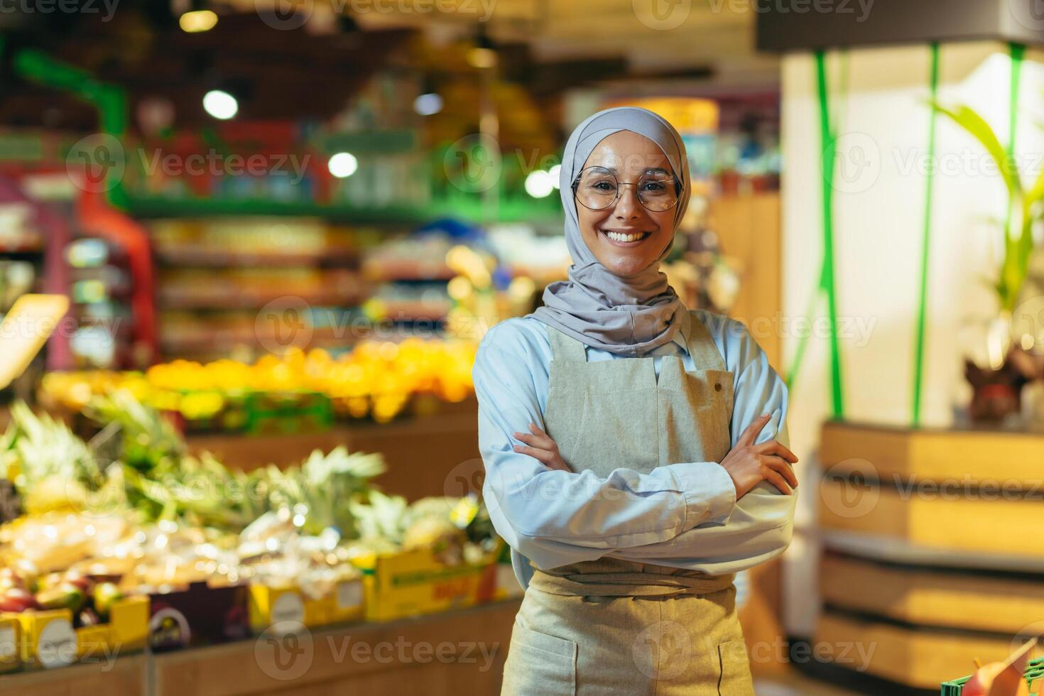 Portrait woman in a hijab as a seller in a supermarket store, a saleswoman with arms crossed smiles and looks at the camera, an apron sells apples and fruits, a Muslim woman is satisfied in glasses photo