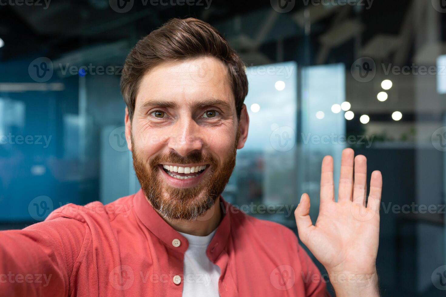 Young happy businessman looking into smartphone camera and talking on call, man working inside modern office building, smiling and talking remotely with friends, using communication app. photo