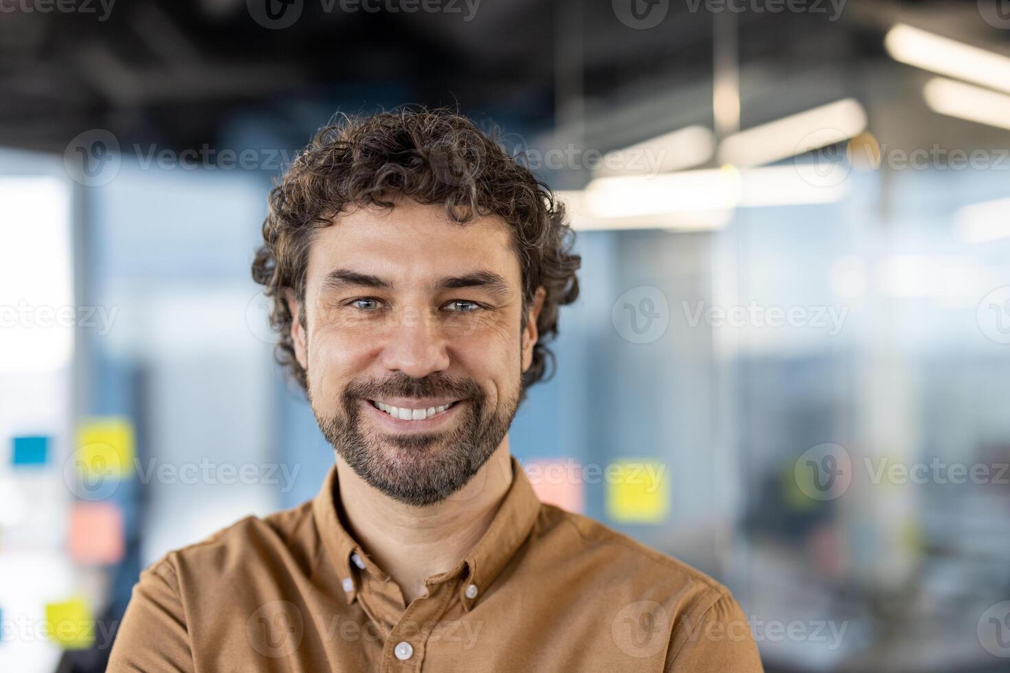 atractivo sin afeitar hombre con Rizado pelo mirando con confianza a cámara con amplio sonrisa en borroso antecedentes. satisfecho empresario gasto ocupado día en empresa edificio y disfrutando trabajando proceso. foto