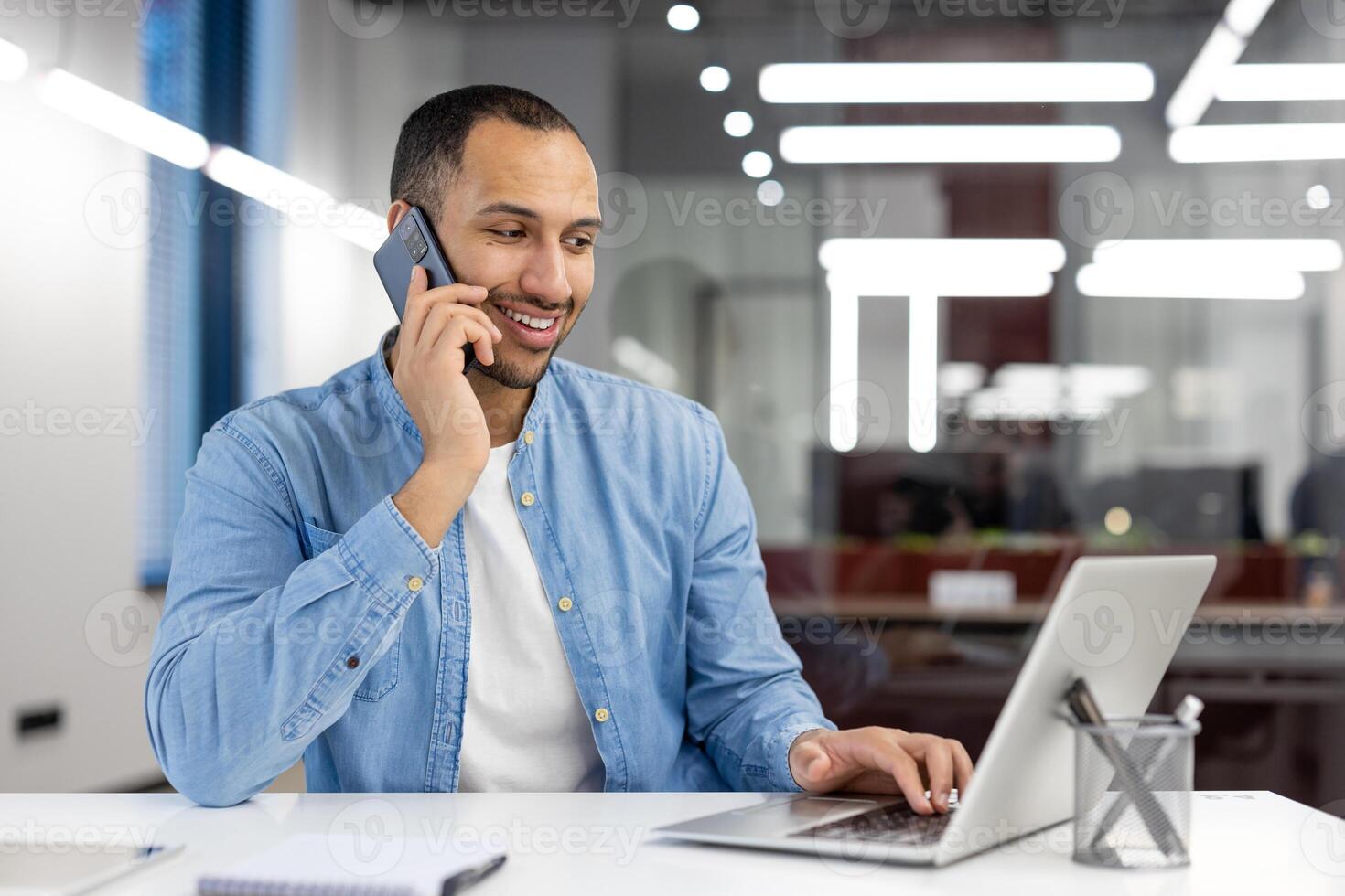 A focused man in a blue shirt multi-tasking in a busy office, talking on the phone while working on his laptop. photo