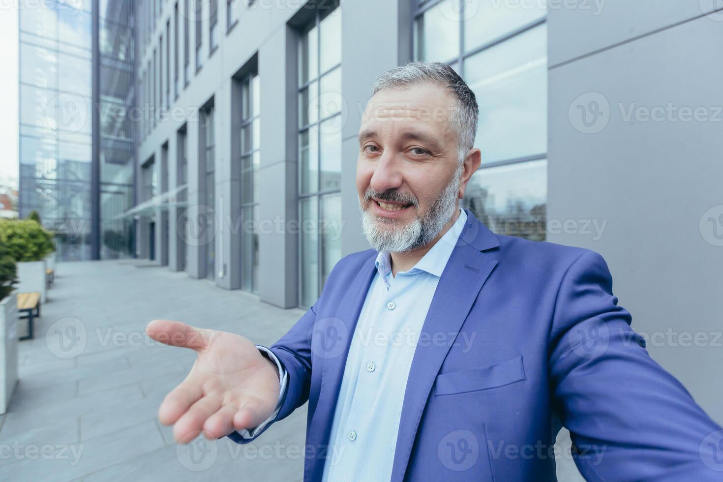 Senior gray-haired handsome man in a suit. Standing near an office building, holding a phone, making a selfie call, talking, pointing, smiling. photo