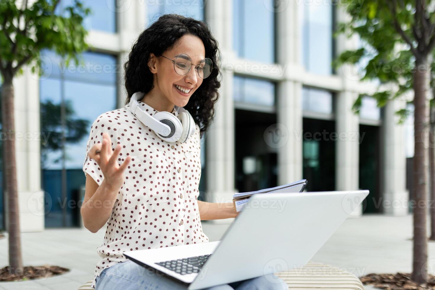 A young female professional engages in a call while working on her laptop outside a modern office building, portraying flexibility and modern work lifestyle. photo
