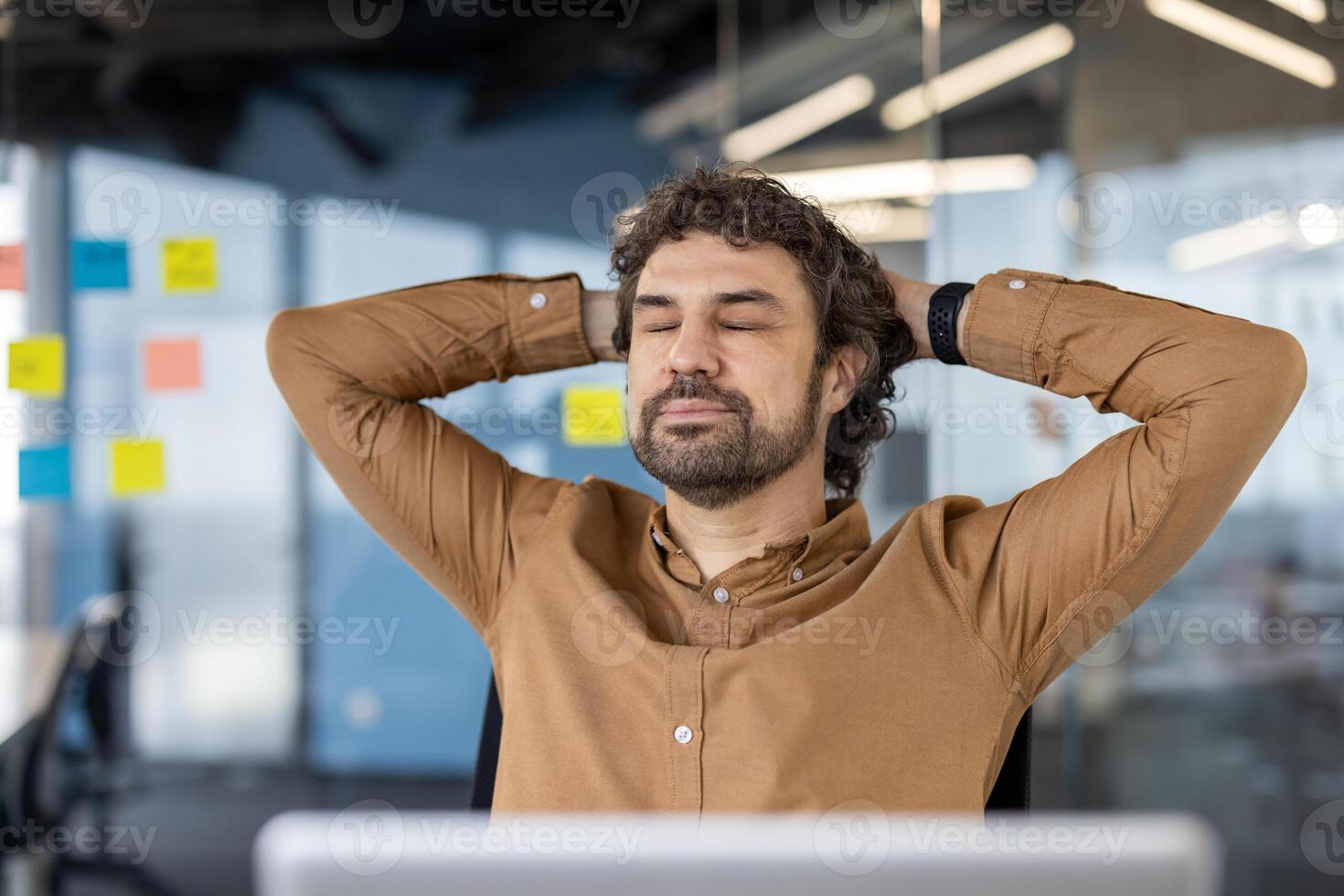 Calm caucasian guy sitting on chair with hands behind head and closed eyes in background of stylish office. Digital designer daydreaming and setting mind up for creative mood at beginning of workday. photo