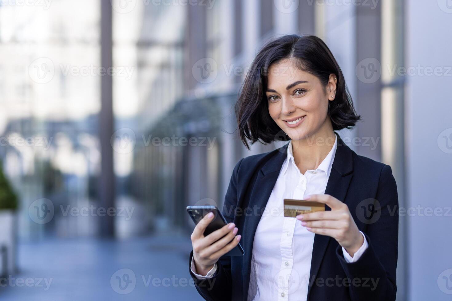 de cerca retrato de un joven negocio mujer en pie cerca un oficina centro, participación un teléfono y un crédito tarjeta, sonriente y mirando a el cámara. foto