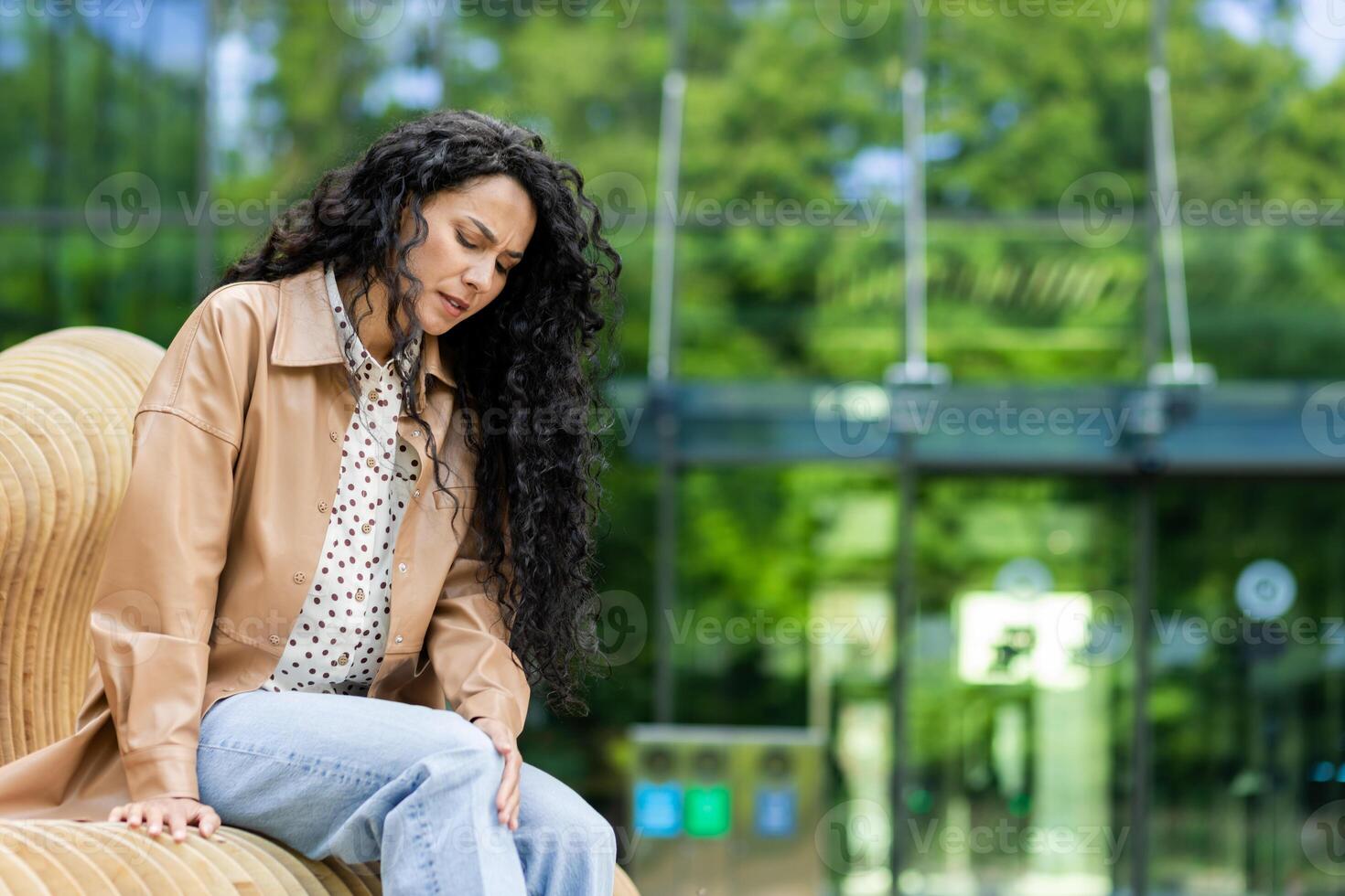 cansado caucásico mujer con largo negro Rizado pelo sentado en banco al aire libre conmovedor y experimentando incomodidad en rodilla. antecedentes de parte de vaso estructura o edificio. foto