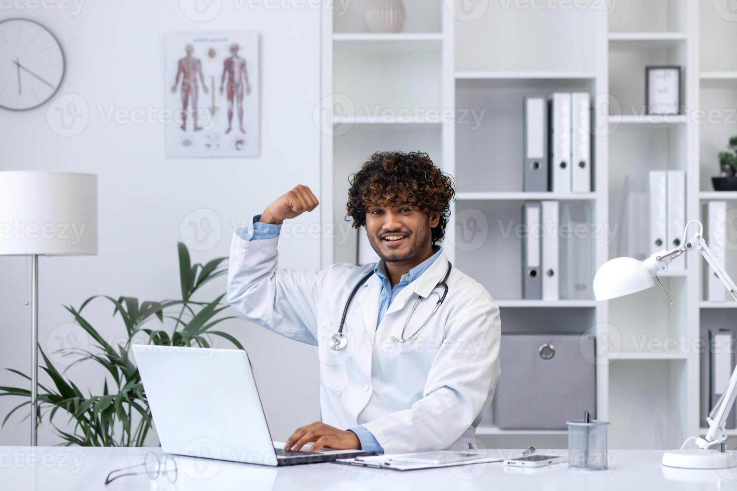 Well done superhero doctor, man smiles looks at camera, holds his hands up, rejoices at the success in the treatment of the patient, works inside the clinic in the medical office, sits at the table. photo