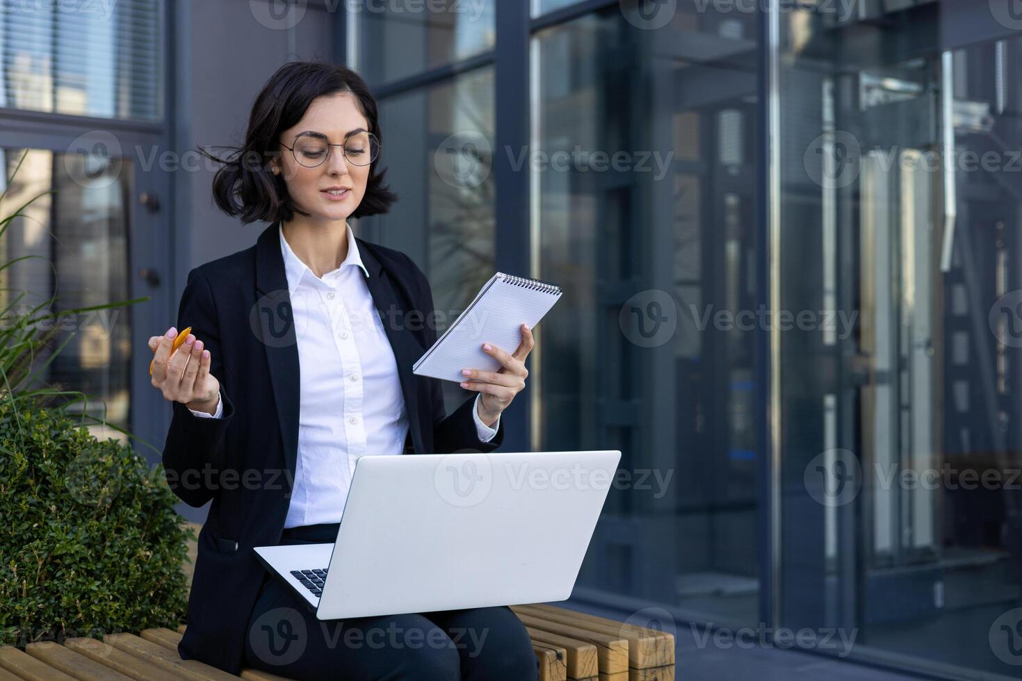 Professional businesswoman in a suit sits outdoors with her laptop and notebook, engaging in work with a focused expression in a modern urban setting. photo