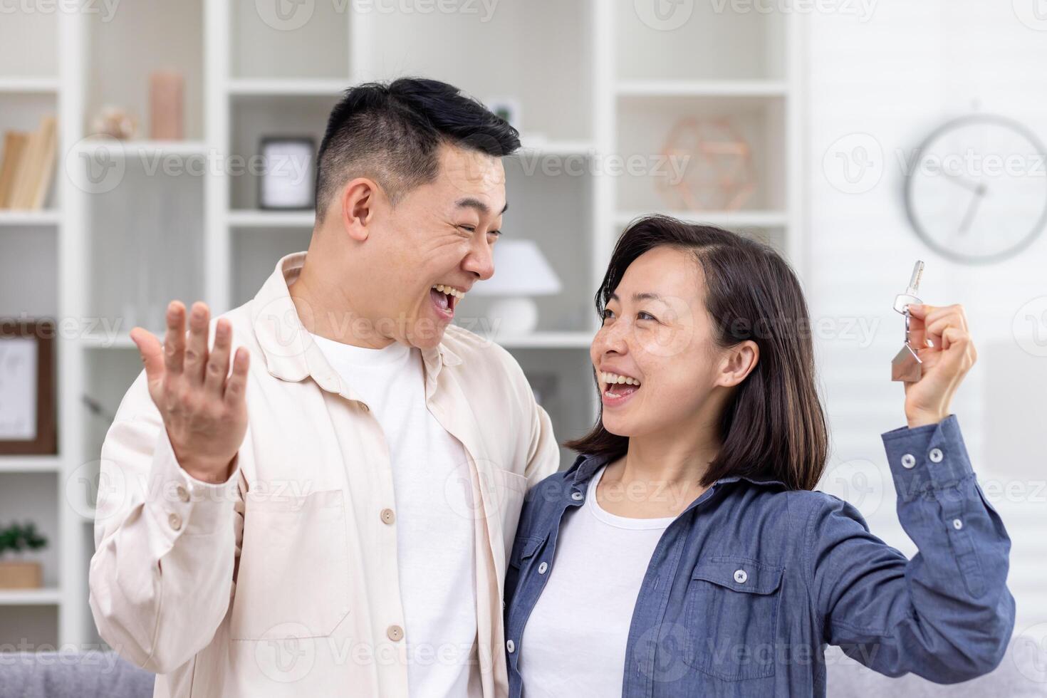 A young Asian couple, a man and a woman, stand hugging in a house, apartment, and hold keys in their hands. Smiling people look at each other, happy buyers, rent. photo