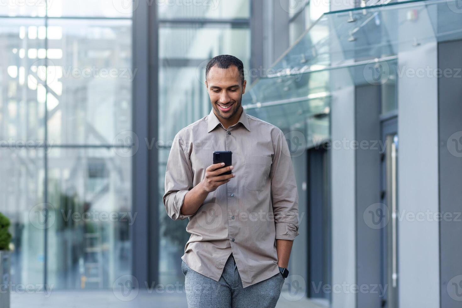 Latin american businessman in casual shirt walking near office building from outside, man smiling and using phone, freelancer typing message and browsing internet pages photo
