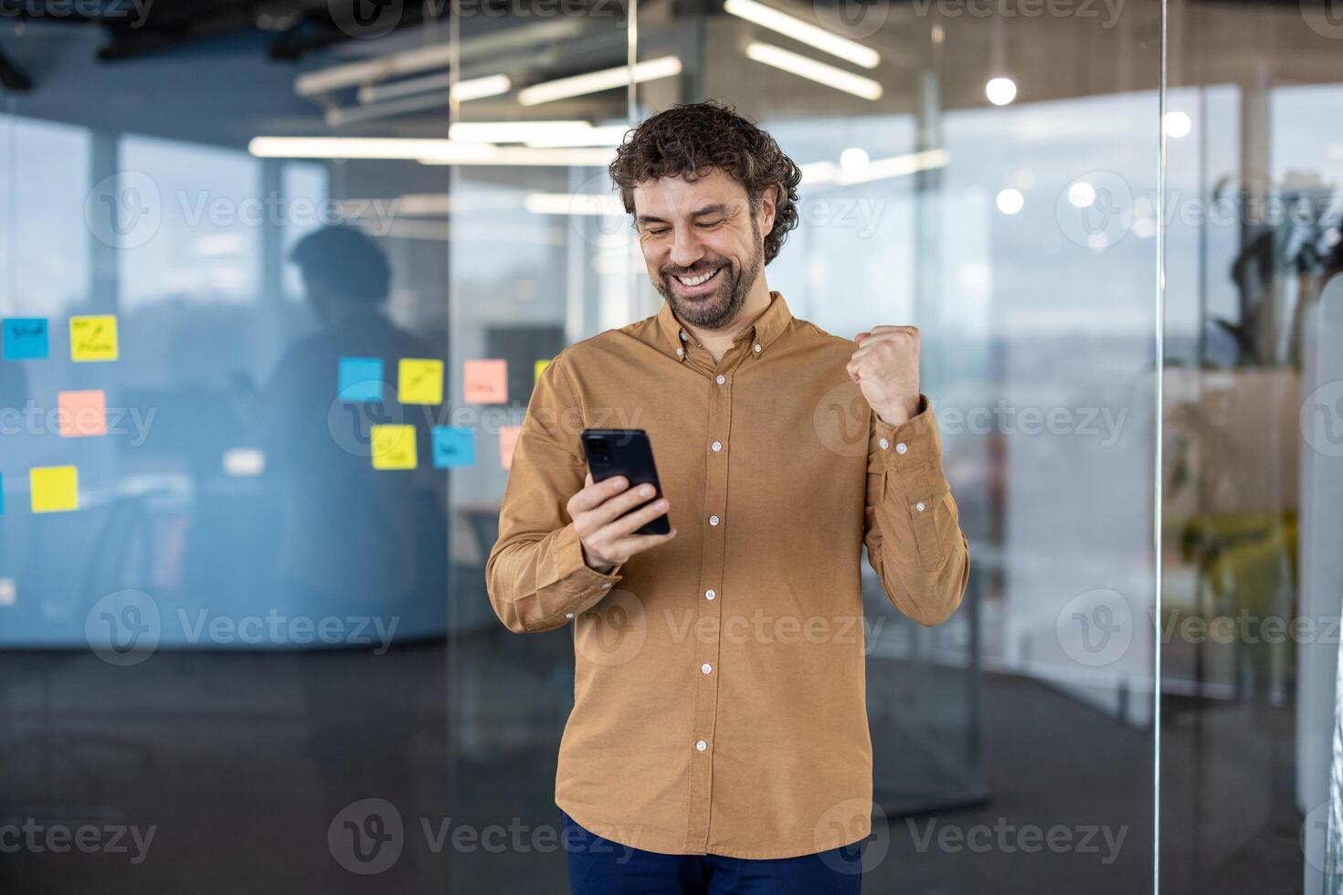 Satisfied millennial male in sandy shirt making victory gesture while looking at screen of mobile phone with happy expression. Lucky guy reading message with good news while going to workplace. photo