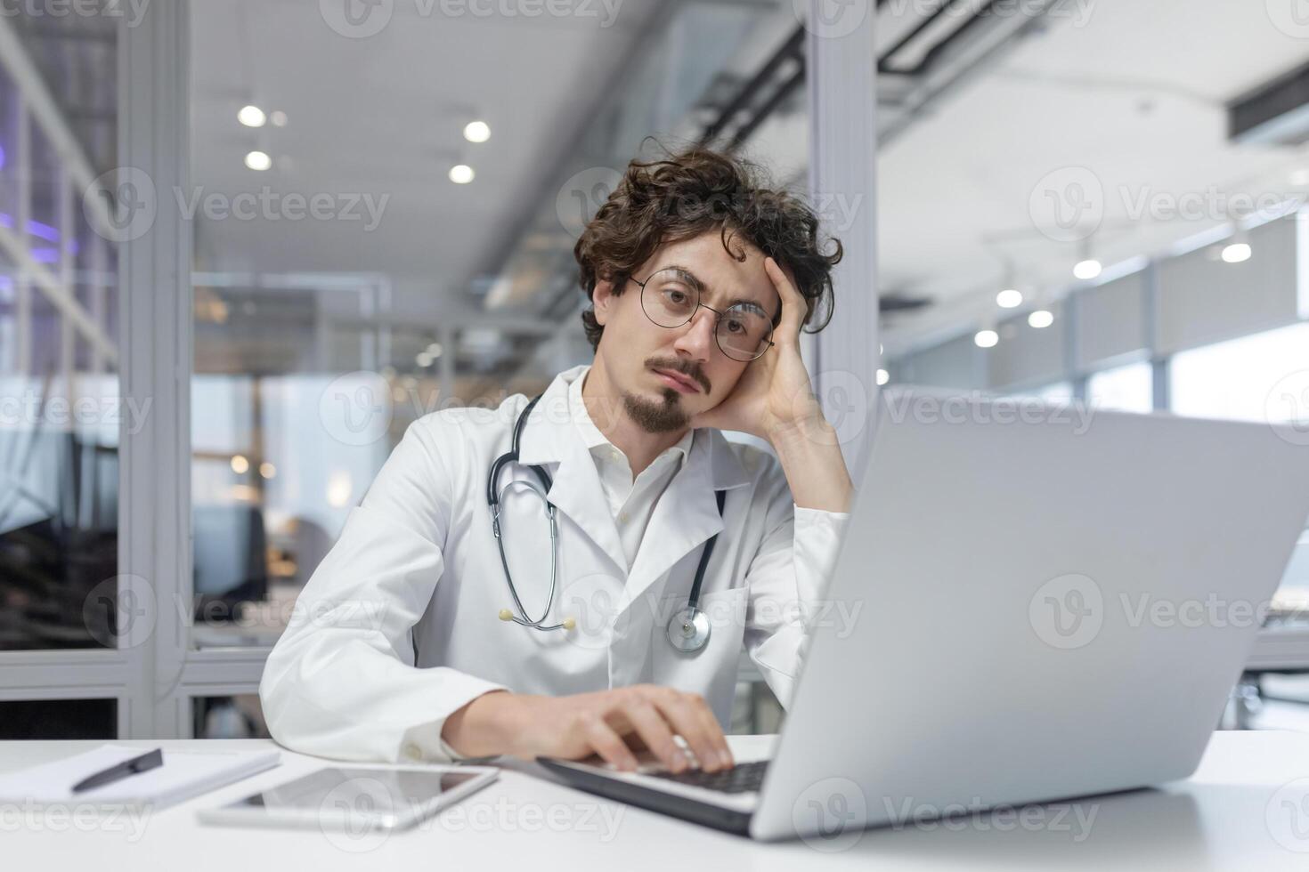 A man in a white medical coat with a stethoscope sits in front of a laptop computer in a medical office. Doctor thinking using computer photo