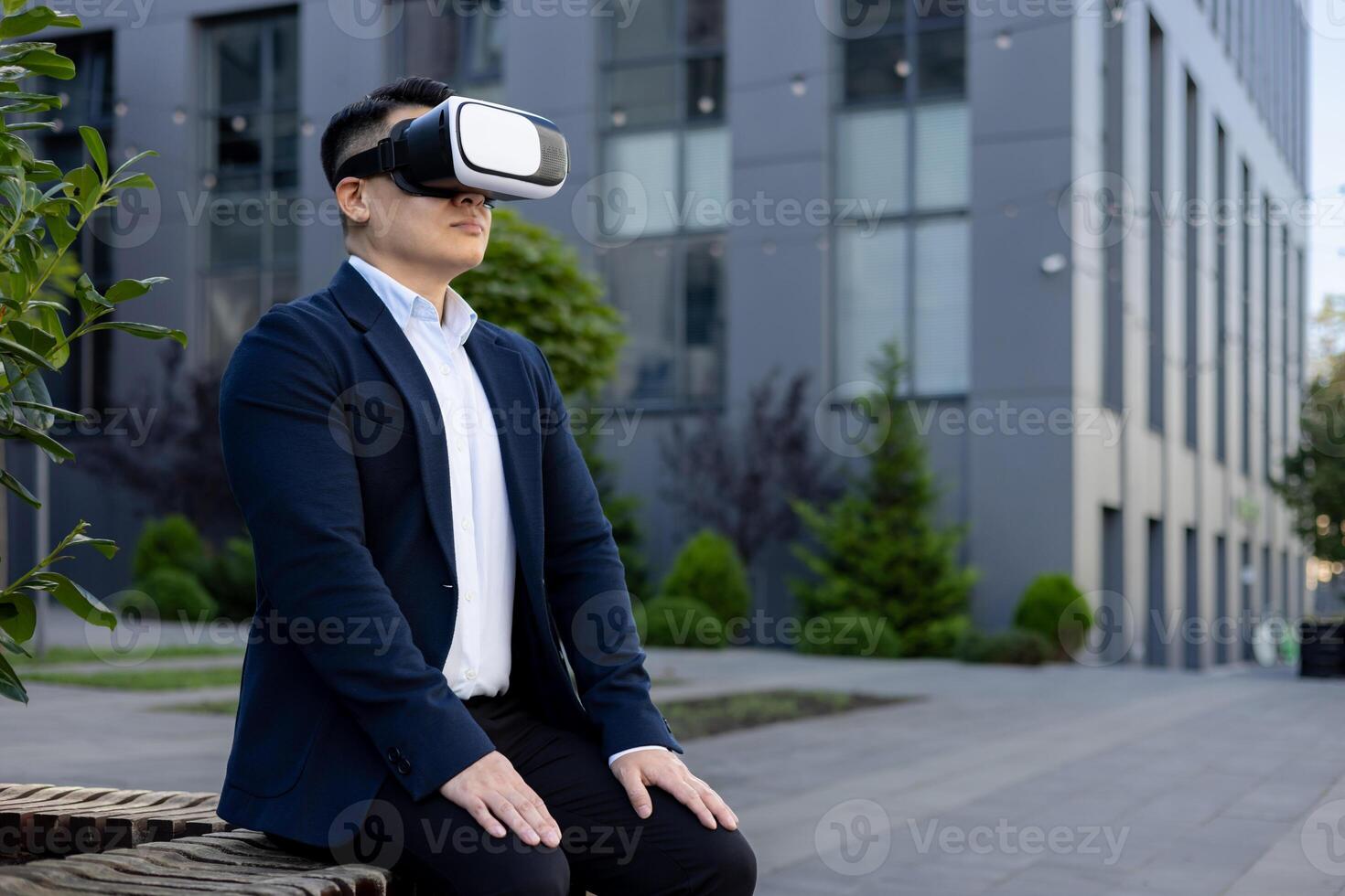 Serious and focused Asian male office worker in a business suit sitting on a bench near a building wearing a virtual mask. photo