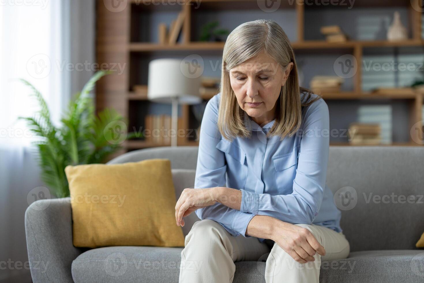 An elderly woman sits alone on her sofa, looking down with a sad expression. The image captures a moment of solitude and emotional struggle often experienced in senior years. photo