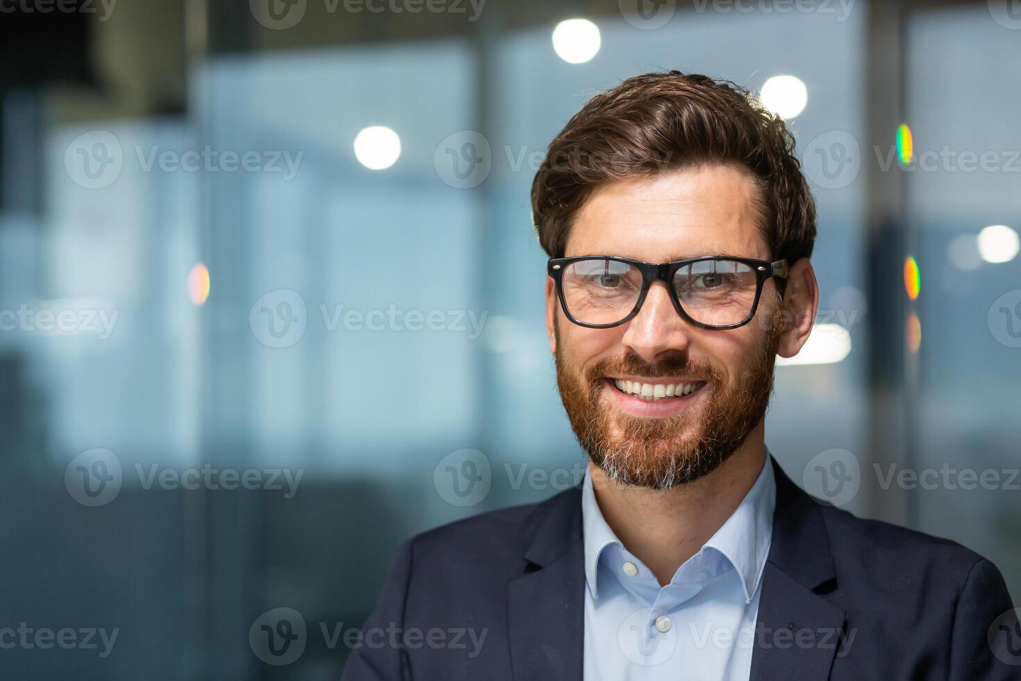 cerca arriba foto retrato de exitoso y contento empresario, maduro jefe con barba y lentes trabajando dentro moderno oficina edificio, mayor inversor sonriente y mirando a cámara.