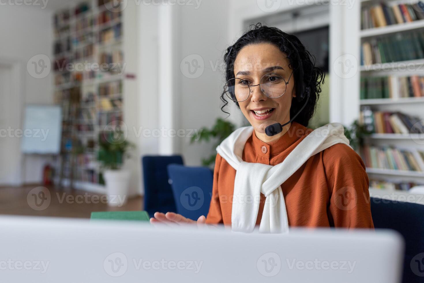 Teacher conducts lessons online, woman with call headset smiling and explaining lecture online using laptop for remote communication and learning, sitting at desk in university library at desk. photo