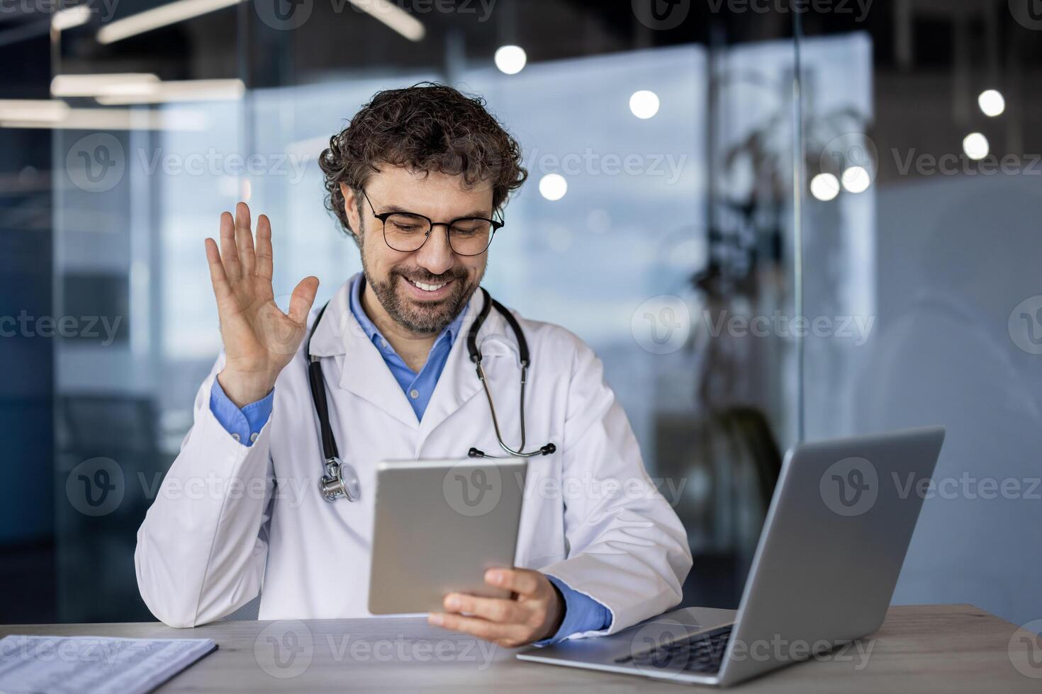 Smiling male doctor in lab coat waving during a telemedicine consultation, expressing approachability and modern healthcare services. photo