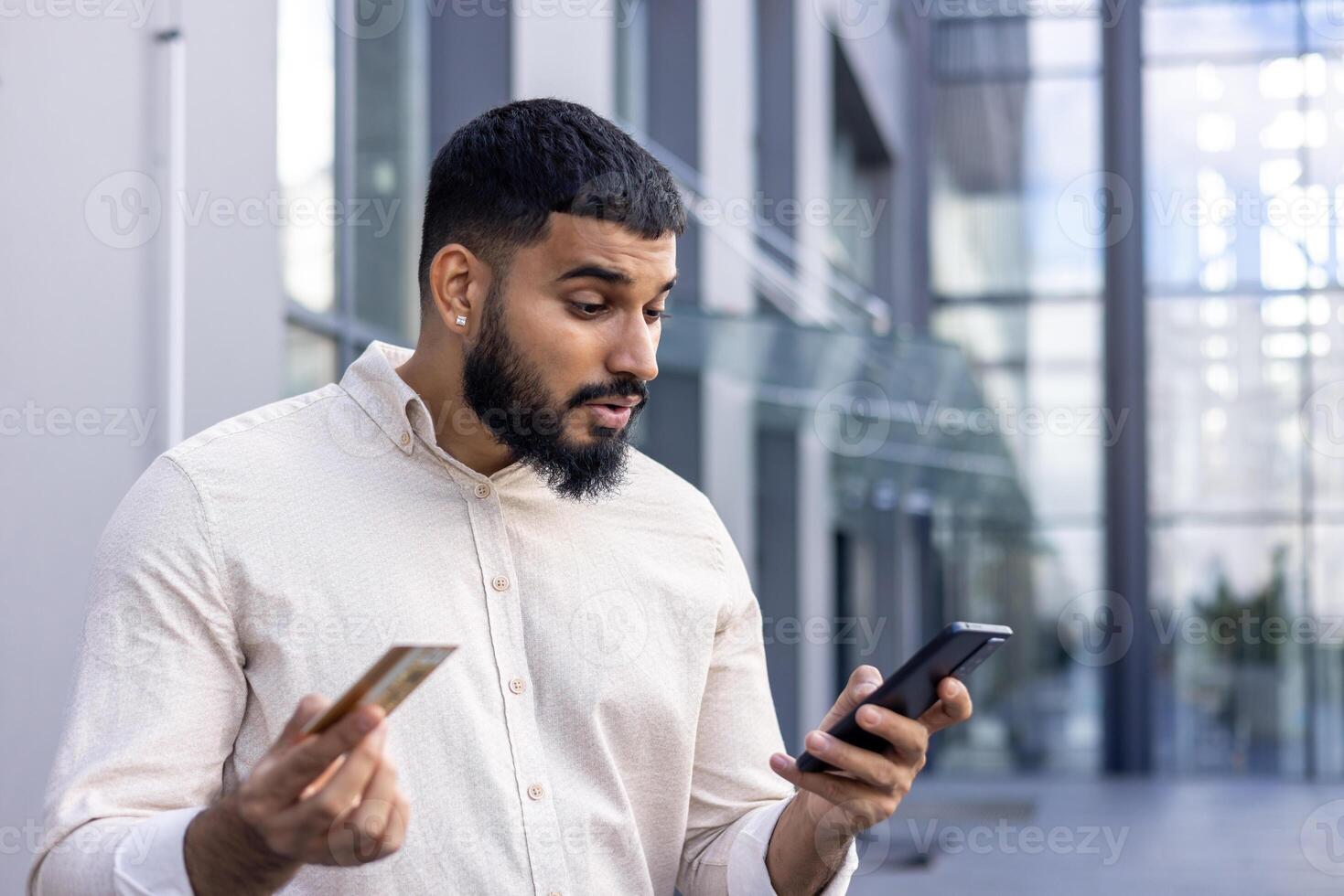 Close-up photo of a sad young Muslim man standing on the street, holding a credit card in his hand and looking disappointedly at the phone screen