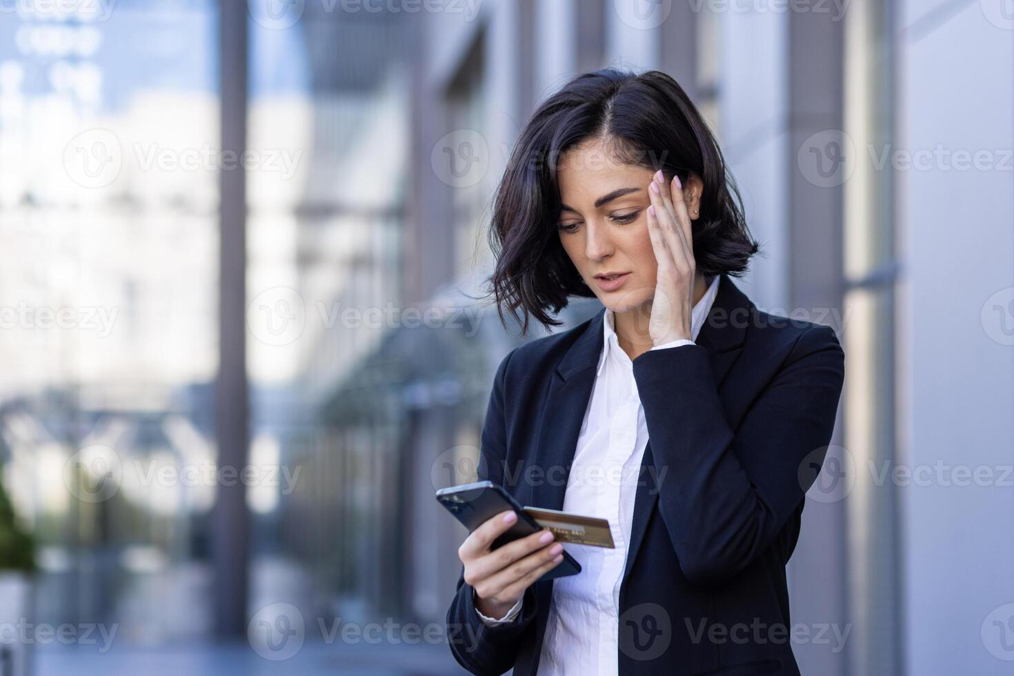 Problems with the financial account. Upset young business woman standing on the street and holding her head, holding a credit card and looking at the phone. photo