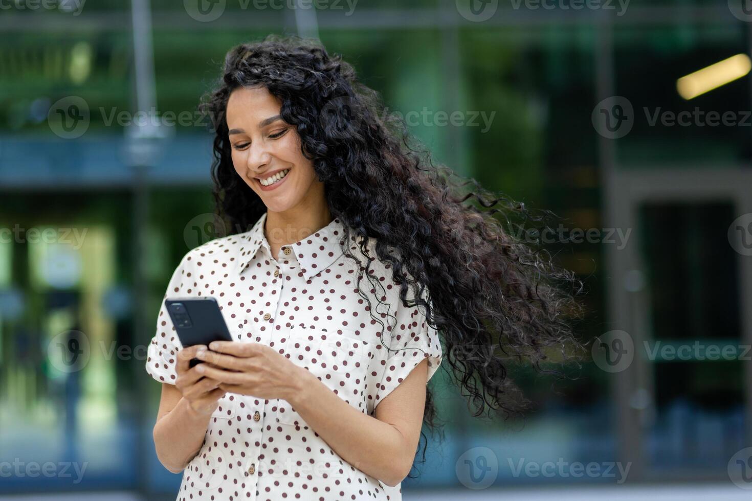 joven hermosa latín americano mujer caminando en el ciudad con teléfono en manos, negocio mujer después trabajando día, sonriente con satisfacción, estudiante utilizando solicitud en teléfono inteligente foto