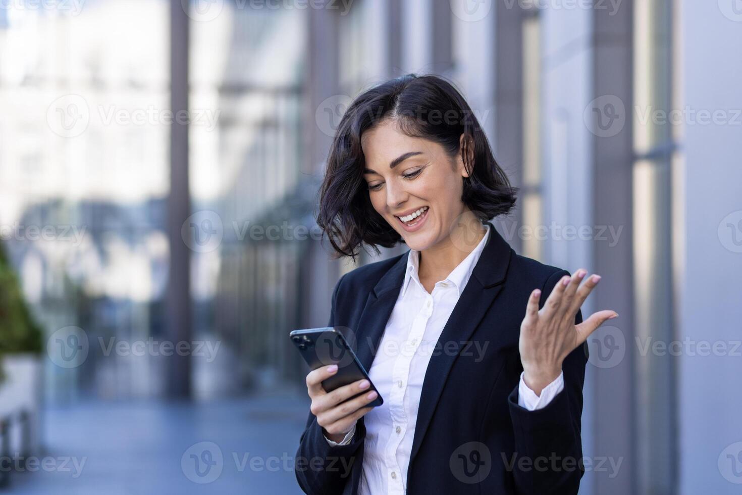 Smiling young businesswoman standing on the street near an office building, using the phone, happy with good news, reading a message. photo