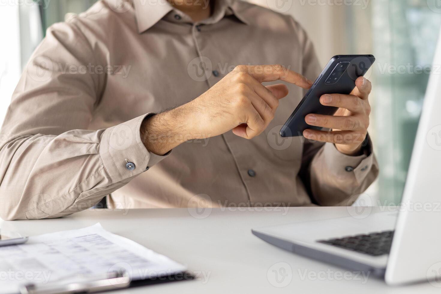 Midsection of a professional man using a smartphone while sitting at a modern office desk with a laptop. Focus on digital communication in a business setting. photo