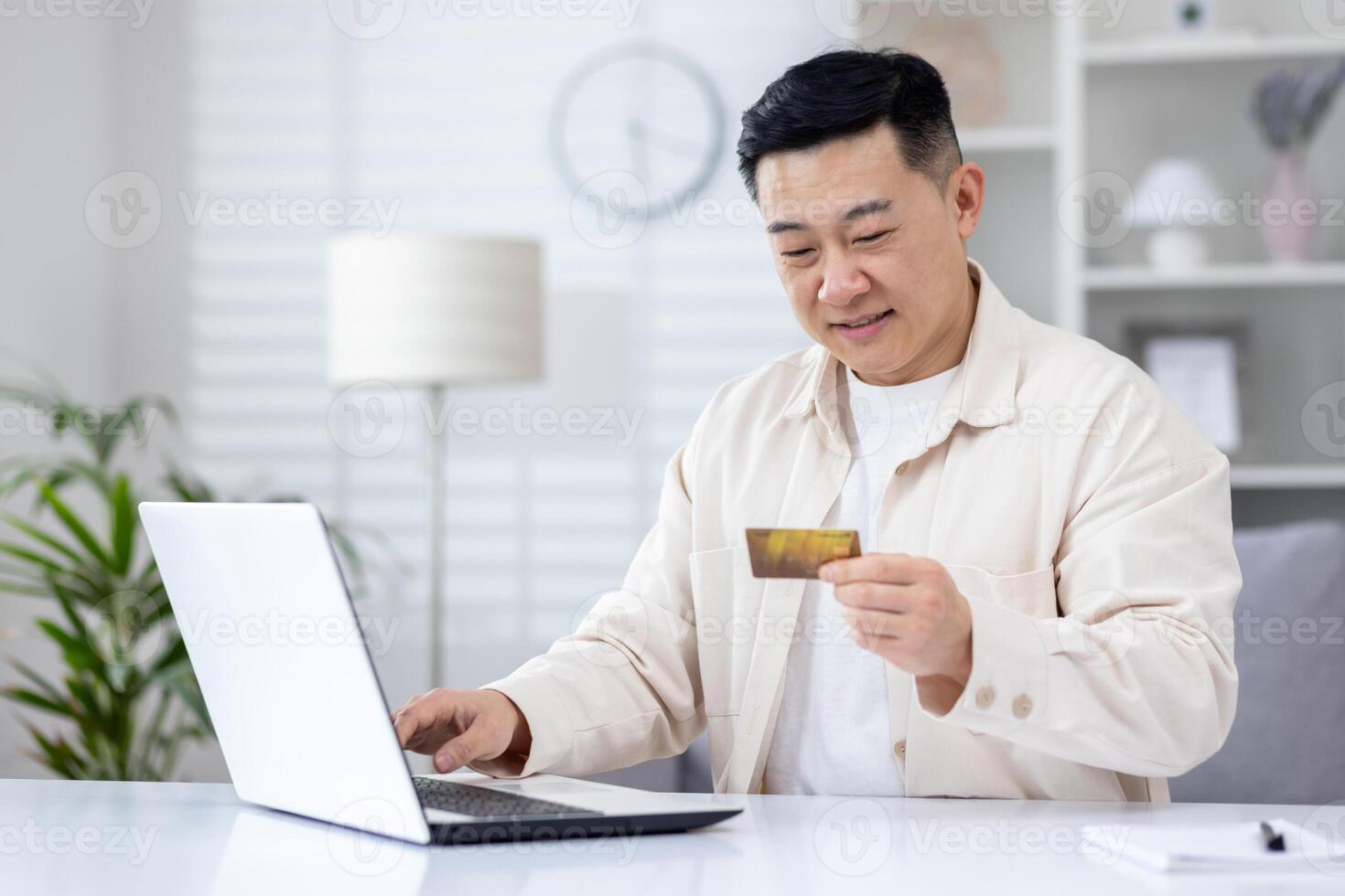 Joyful successful asian businessman working from bright home office, man using bank credit card and laptop for online shopping in online store, and booking services remotely. photo