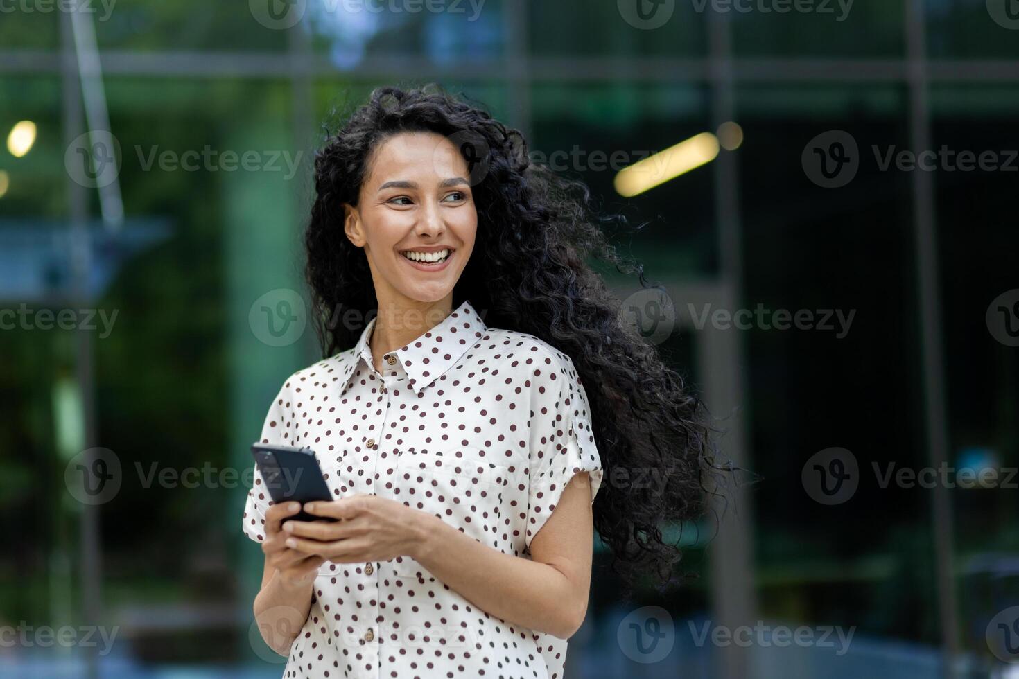 Young beautiful Latin American woman walking in the city with phone in hands, business woman after working day, smiling with satisfaction, student using application on smartphone. photo