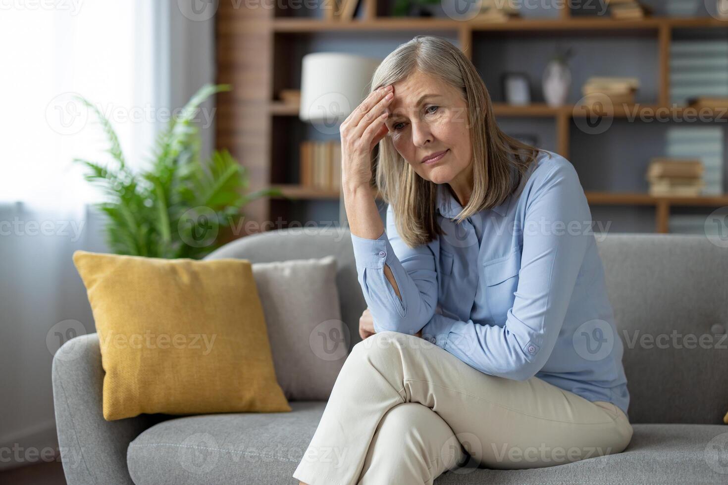 Thoughtful elderly lady sitting cross-legged and resting head on hand with frustrated expression.Tired woman suffering from headache while rubbing forehead and looking forward with disappointment. photo