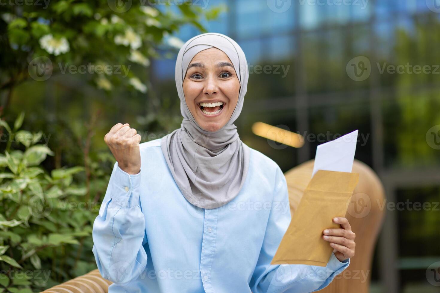 Vibrant image of a joyful Muslim woman wearing a hijab, exuberantly celebrating while holding an envelope outdoors with a blurred green background. photo