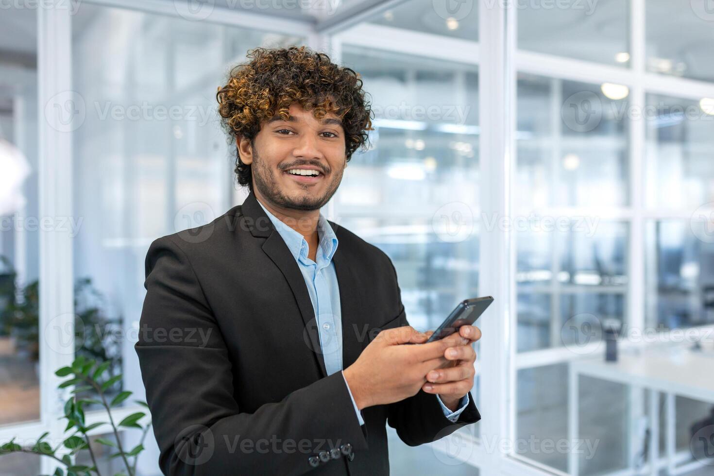 retrato de exitoso Hispano empresario inversor, hombre sonriente y mirando a cámara, jefe en traje a trabajo dentro oficina participación teléfono inteligente foto