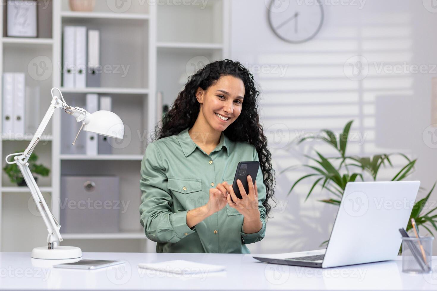 Professional hispanic woman working in a bright office space with a laptop and mobile phone, displaying efficiency and multitasking skills. photo