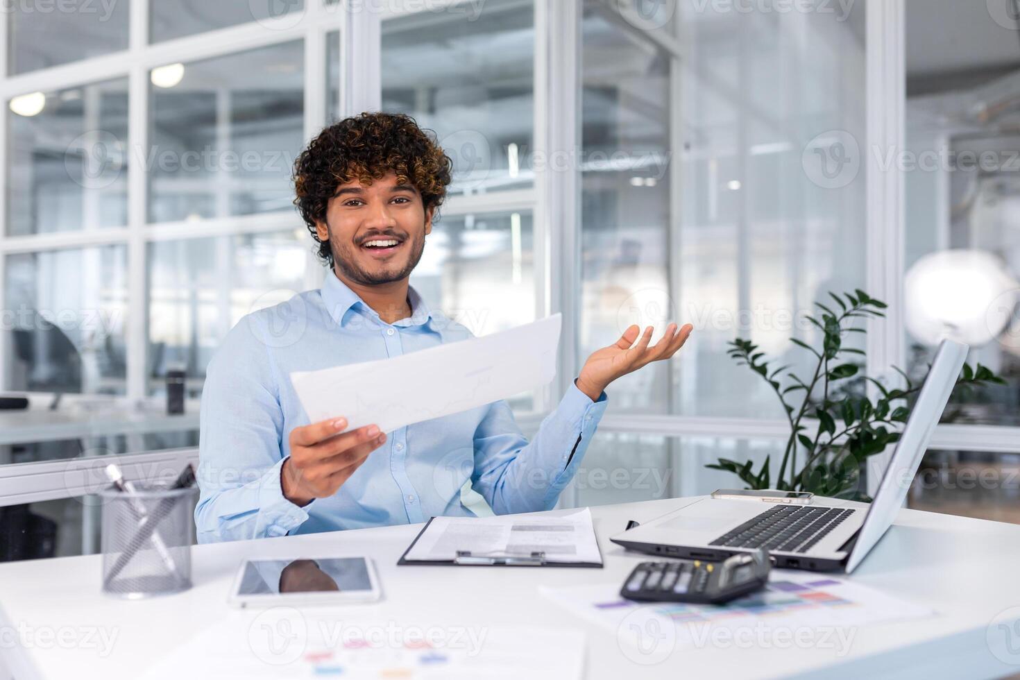 A young African-American male designer, architect sitting in the office at the table with a laptop. He holds documents in his hands, working with plans and drawings, looks at the camera, smilling. photo