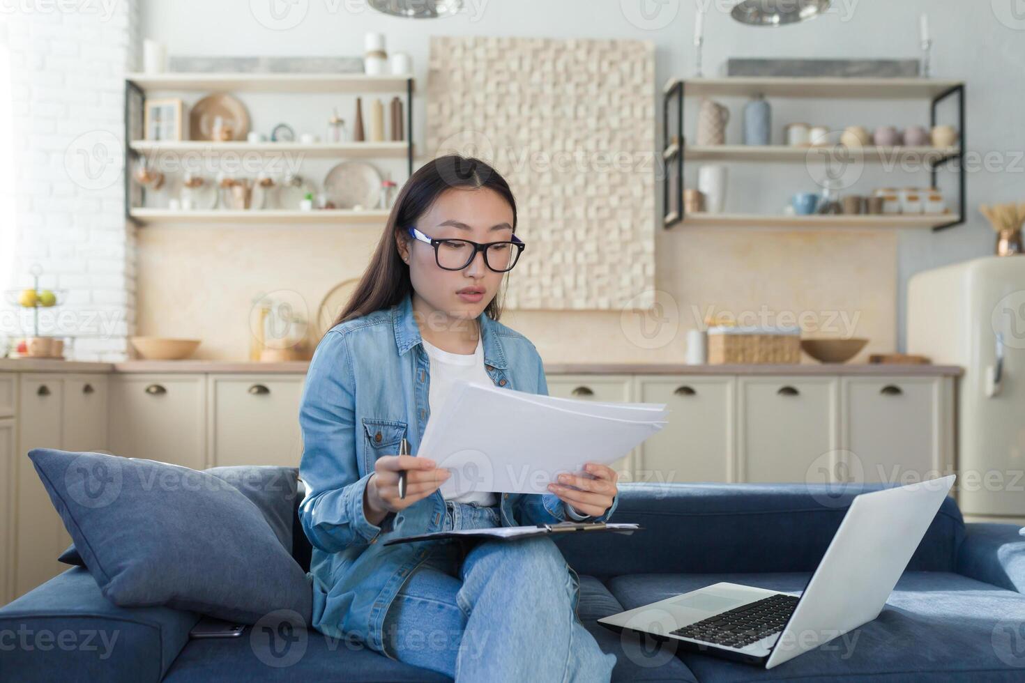 Young beautiful creative woman Asian writer, journalist. He sits at home on the sofa with a laptop and writes a book, an essay, a script, an article. Holds paper and pen. photo