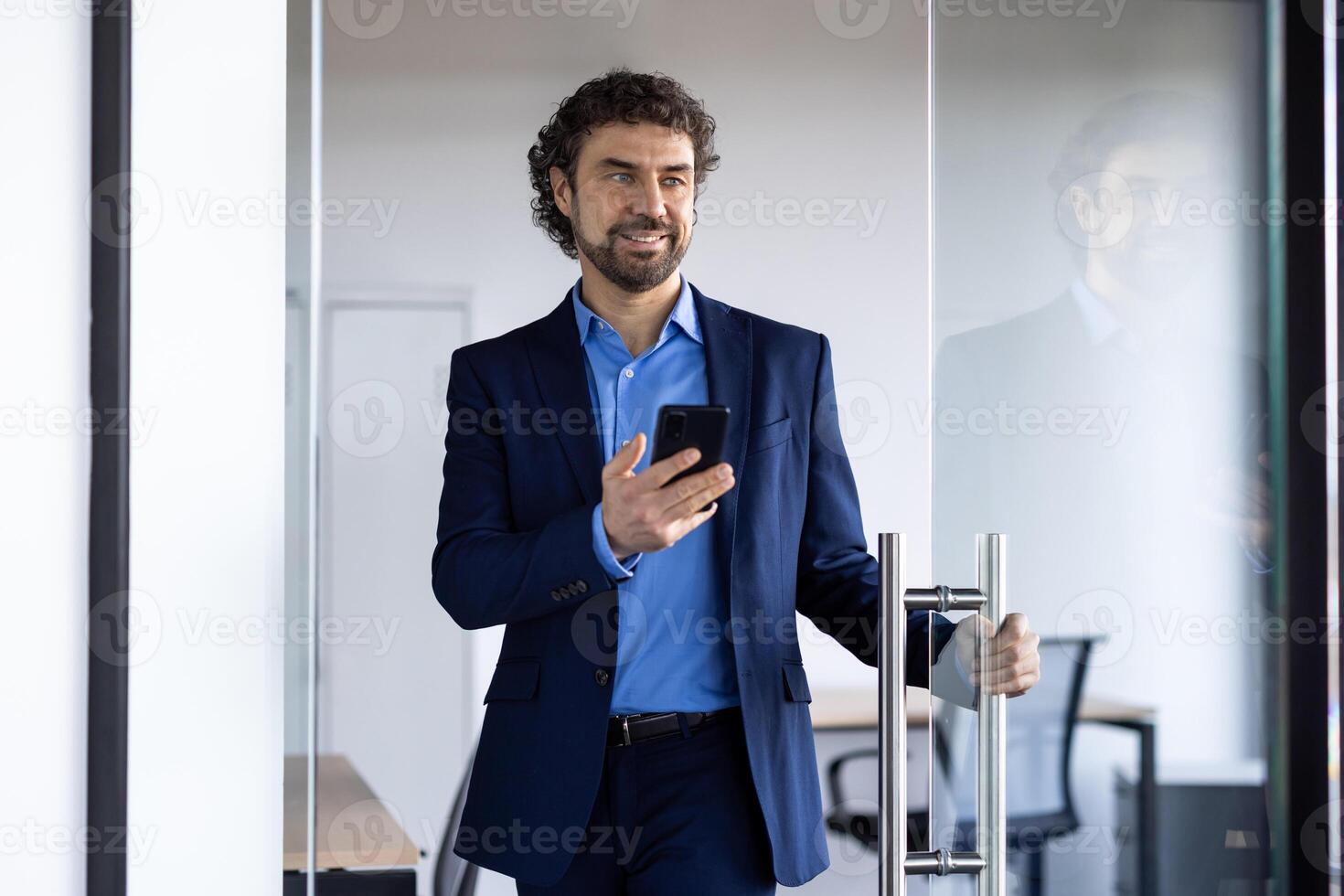 Successful joyful businessman opens glass office door, man with phone in hands smiling, satisfied with achievement results, boss in business suit. photo