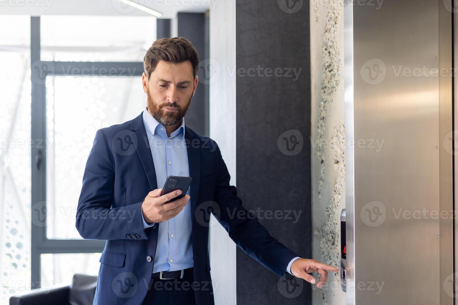 Focused businessman in a suit using smartphone while waiting for an elevator in a modern office building interior. photo