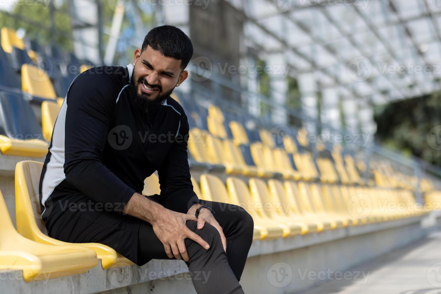A male athlete in workout attire experiencing knee pain while sitting on yellow stadium seats, highlighting sports injury and fitness concepts. photo