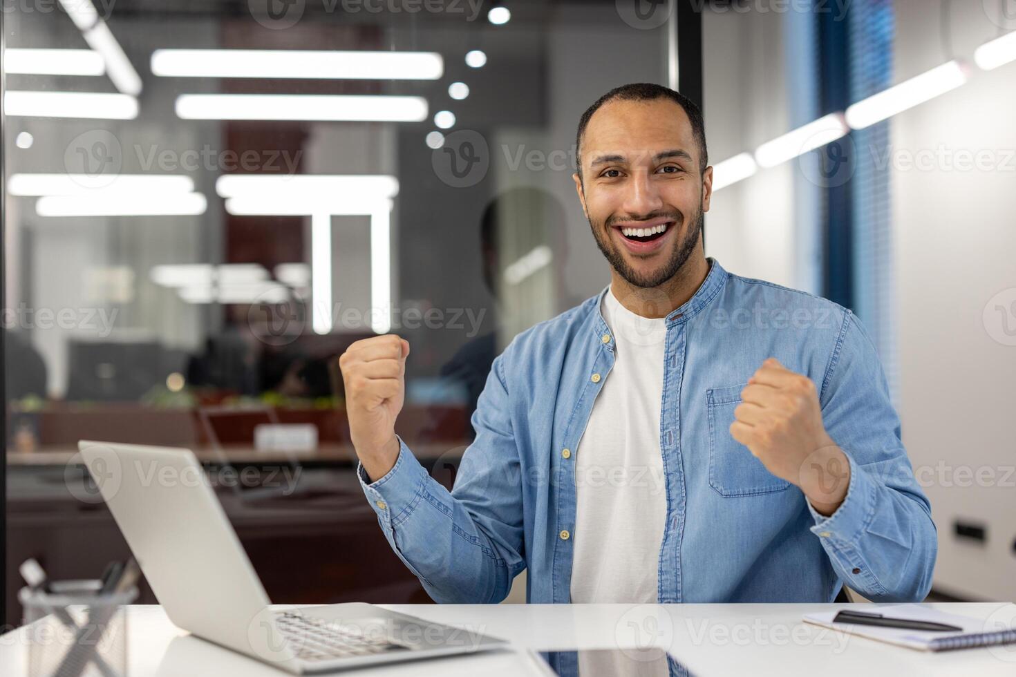retrato de un contento y satisfecho joven musulmán hombre sentado en el oficina a el mesa con un ordenador portátil y alegría en éxito, mirando a el cámara y demostración un victoria gesto con dos manos. foto